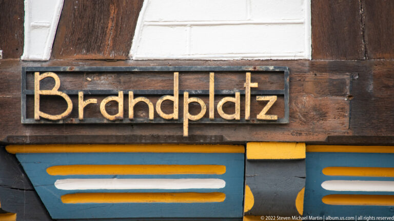 Gold “Brauplatz” lettering on a dark wooden sign mounted to a brown-and-white half-timbered facade.