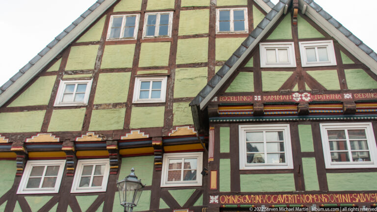 Adjoining green and burgundy half-timbered houses with painted inscriptions, white windows, steep gables, and a street lamp.