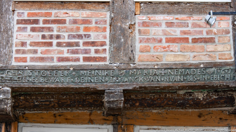 Close view of a carved German inscription in a dark timber beam beneath brick infill on a half-timbered building.