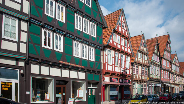 Colorful row of gabled half-timbered houses receding along a Celle street with ground-floor shops and parked vehicles.