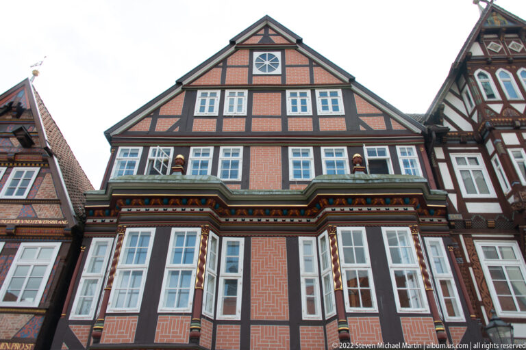 Half-timbered townhouse with patterned brick infill, projecting window bay, circular gable opening, and steep roof.
