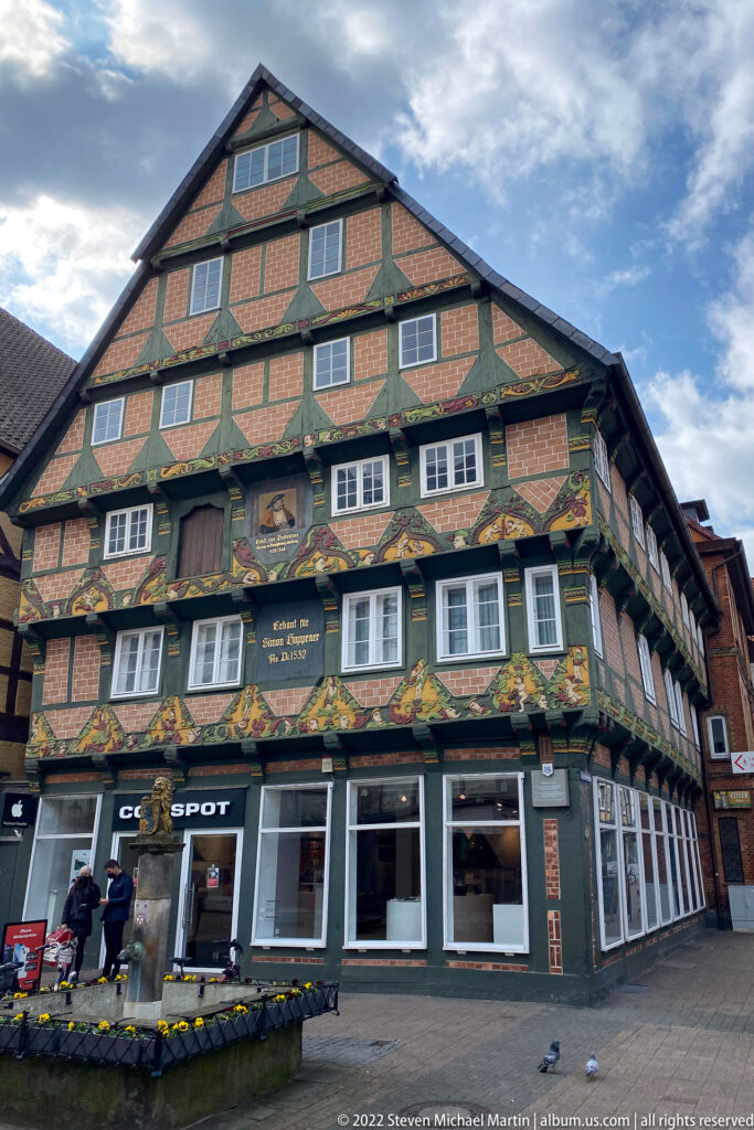 Large half-timbered corner house with brick infill, painted friezes, projecting floors, rows of windows, and shops below.