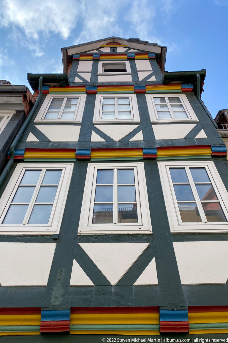 Low-angle view of black-and-white half-timbered framing with colored horizontal trim and white-framed windows under a gable.