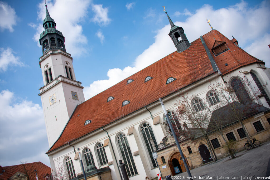 Pale church with tall Gothic-style windows, steep red tile roof, and square tower topped by a green copper lantern.