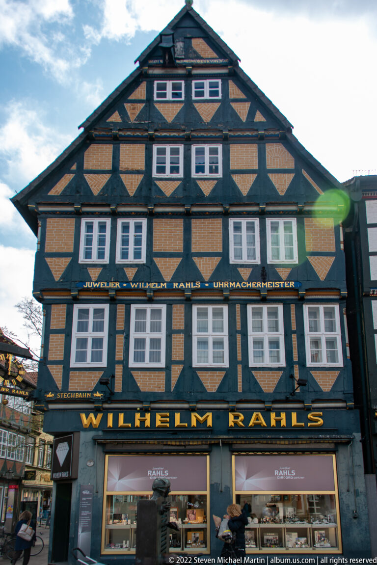 Broad half-timbered building with ochre infill, rows of white windows, and large gold “WILHELM RÄHLS” storefront lettering.