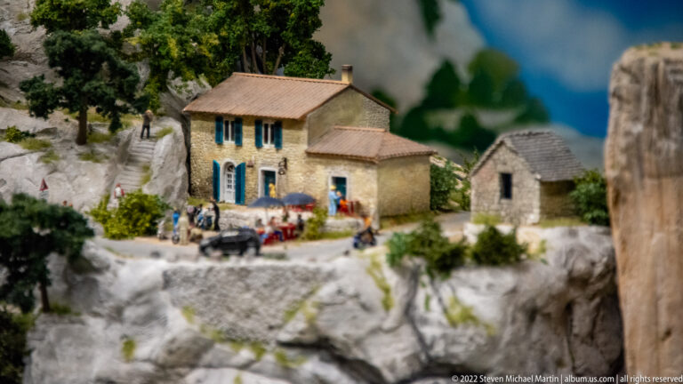 Miniature two-story stone house with blue shutters, parked vehicles, figures, and a rocky cliff setting.