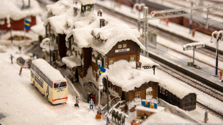 Snow-covered miniature Kiruna station beside railway tracks, with a bus and small passengers near the entrance.