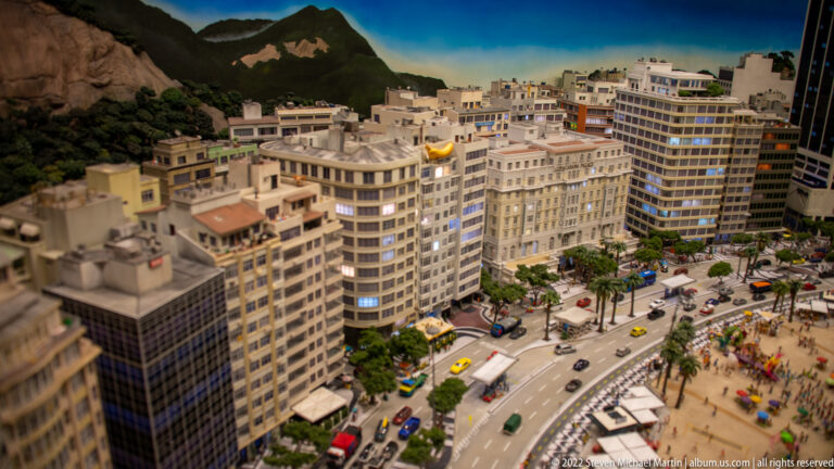 Miniature beachfront city with high-rise apartments, palms, traffic, sandy shore, and mountains in the background.