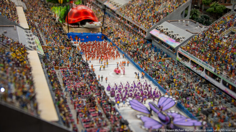 Colorful miniature samba parade moves through a packed grandstand-lined avenue with purple and red performers.