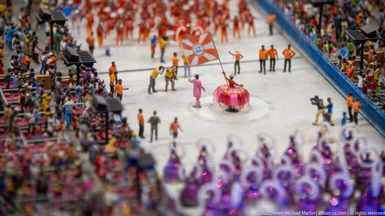 Close view of miniature samba parade performers, purple-costumed dancers, and packed spectators along the route.