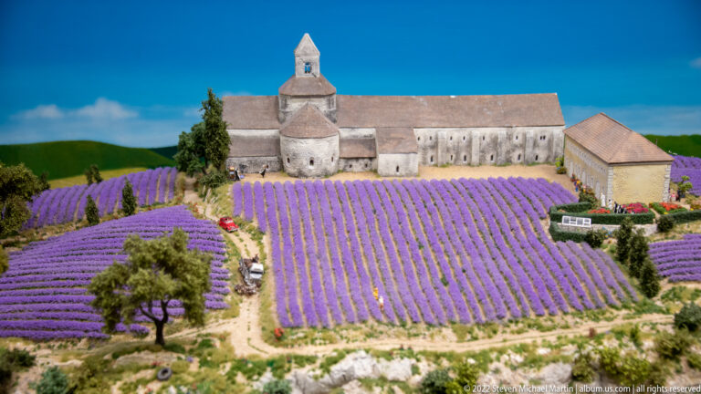Miniature stone abbey behind broad rows of purple lavender under a blue sky backdrop.