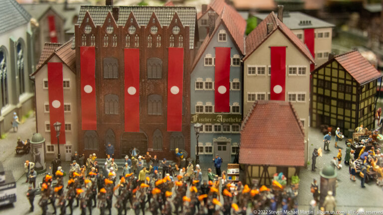 Miniature orange-clad procession passes gabled European buildings decorated with tall red banners.