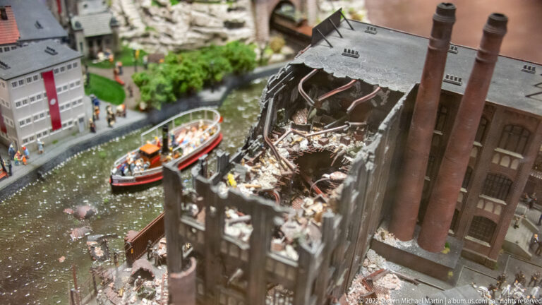 Collapsed miniature factory with exposed rubble and smokestacks beside a canal carrying tour boats.