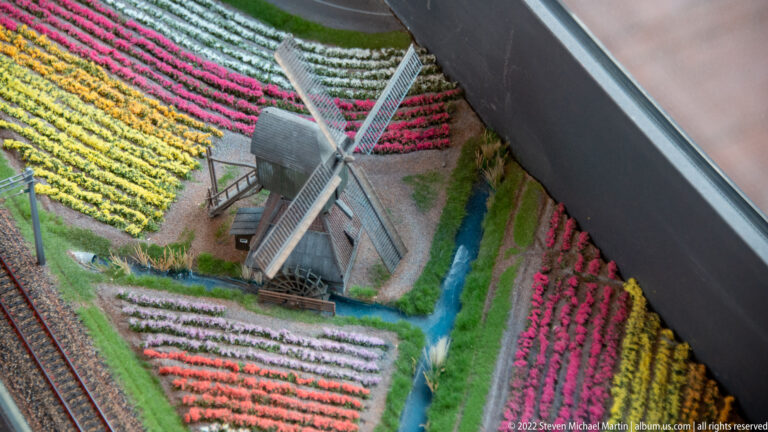 Traditional miniature windmill surrounded by striped tulip fields, canals, and paths.