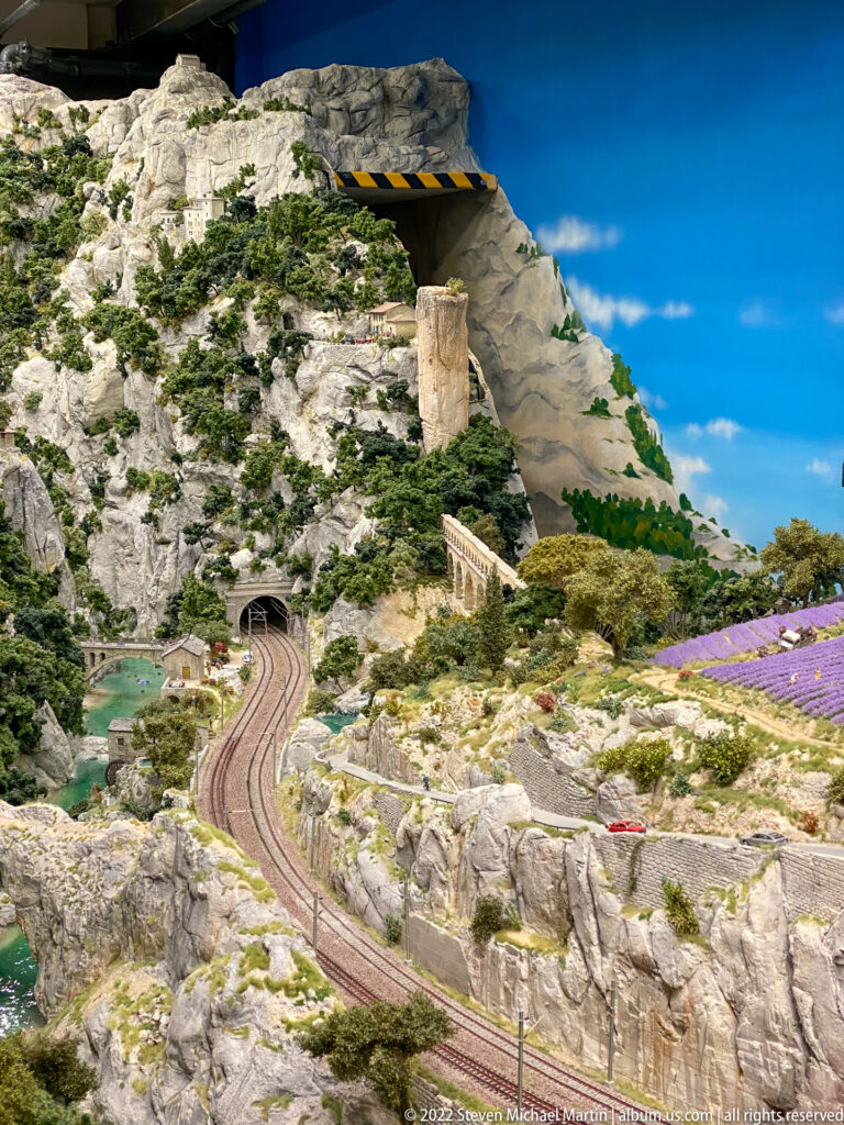 Double-track miniature railway curves through rocky alpine terrain with lakes, tunnel portal, trees, and purple fields.
