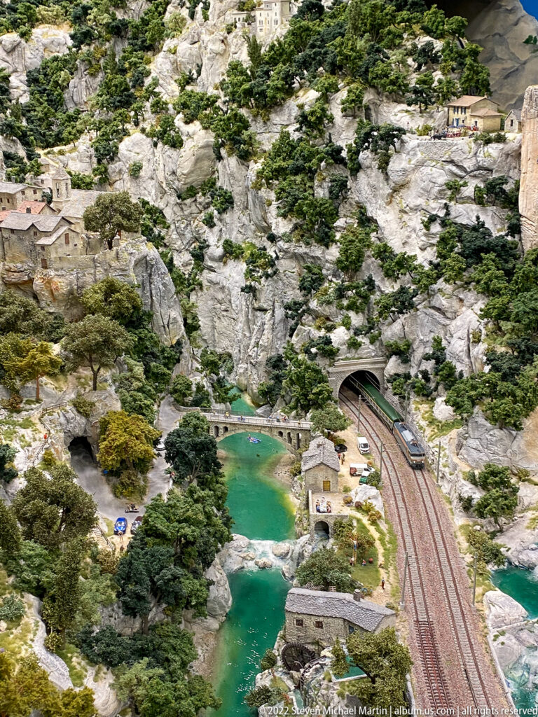 Miniature railway tracks enter a mountain tunnel beside turquoise water, stone buildings, trees, and rocky slopes.
