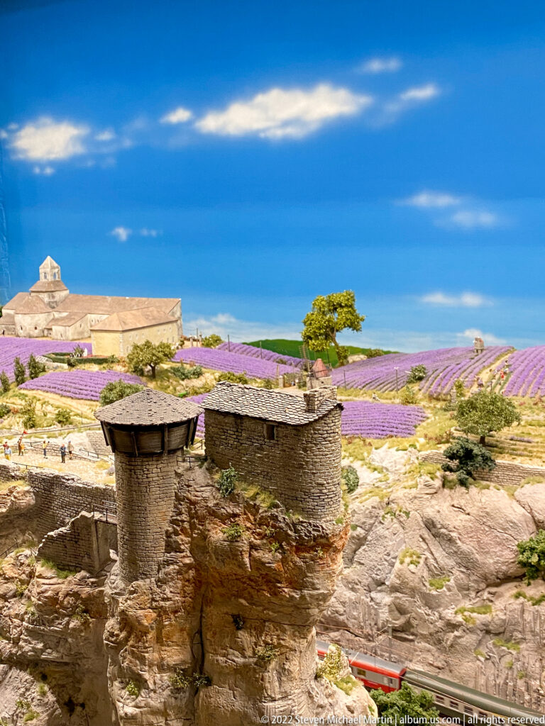 Miniature lavender fields extend behind rough stone huts, rocky ground, and a solitary tree under a blue sky.