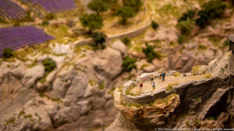 Miniature visitors on a curved stone overlook above steep rocky cliffs and a gorge path.