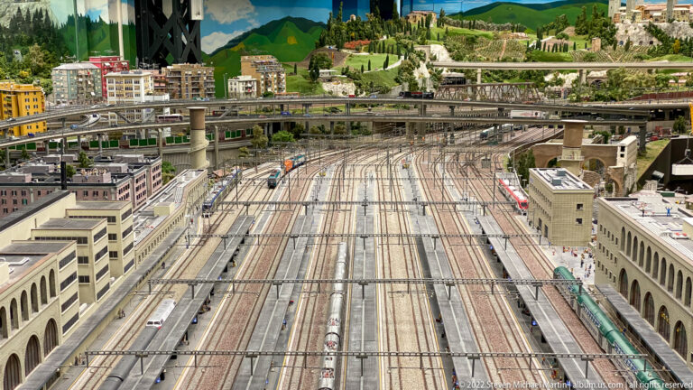 Large miniature electrified rail yard with many tracks, catenary, platforms, trains, and elevated roadways.