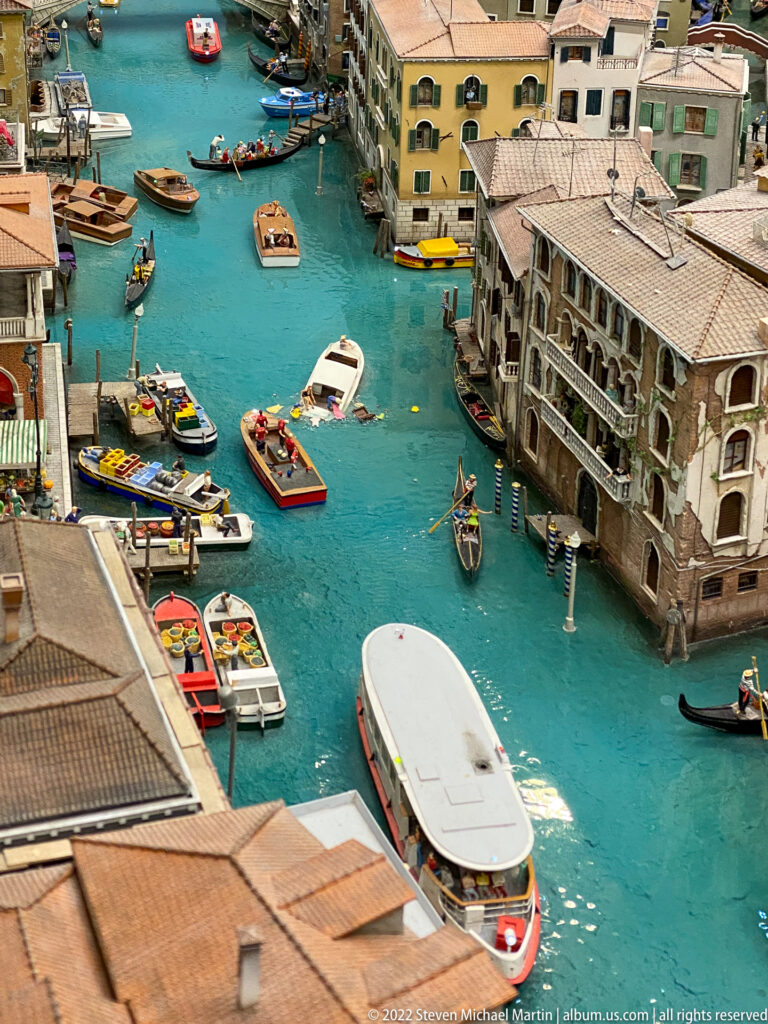 Miniature Venetian canal with gondolas, small boats, a large white waterbus, and palazzo façades.