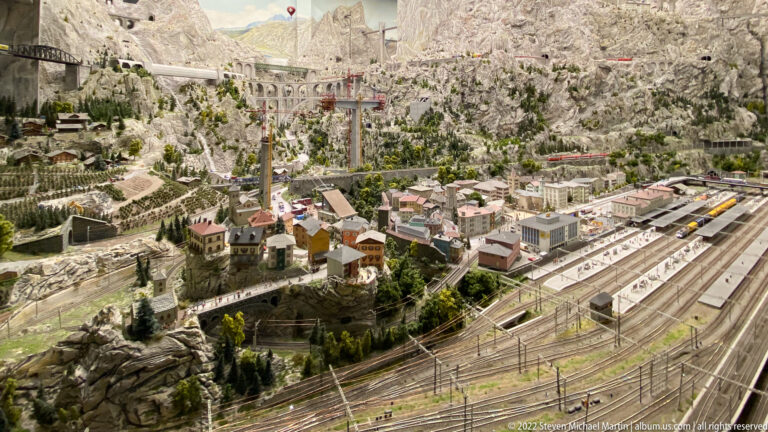 Miniature alpine town with railway station, multiple tracks, cableway structures, roads, and steep rocky mountains.