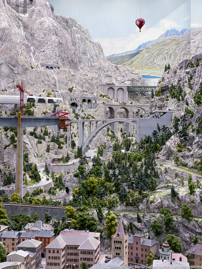 Miniature alpine valley with concrete viaducts, tunnels, cableway towers, red gondola, conifers, and distant lake.