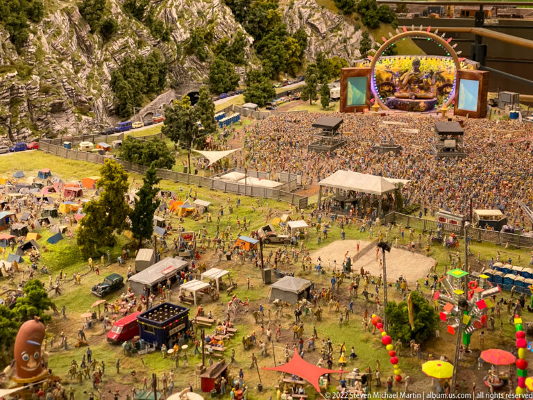 Large miniature outdoor music festival with main stage, dense crowds, tents, temporary structures, and surrounding campground.
