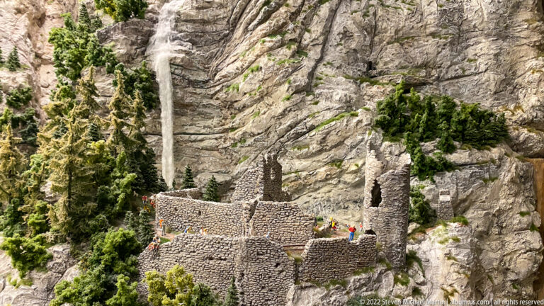 Miniature stone castle ruins on a rocky ledge beneath a steep mountain cliff, with conifers and tiny figures.