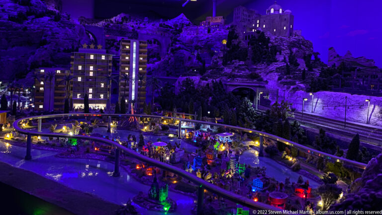 Purple-lit miniature mountain town at night with elevated railway, tunnels, glowing windows, and colorful activity below.