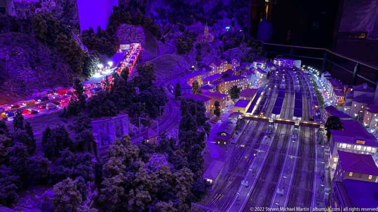 Large miniature railway station and rail yard at night with parallel tracks, platforms, buildings, and blue-purple lighting.