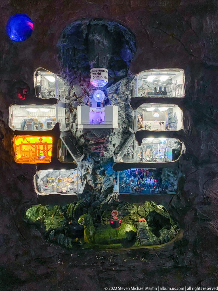 Cutaway miniature underground facility with stacked illuminated rooms, tiny figures, and equipment embedded in dark rock.