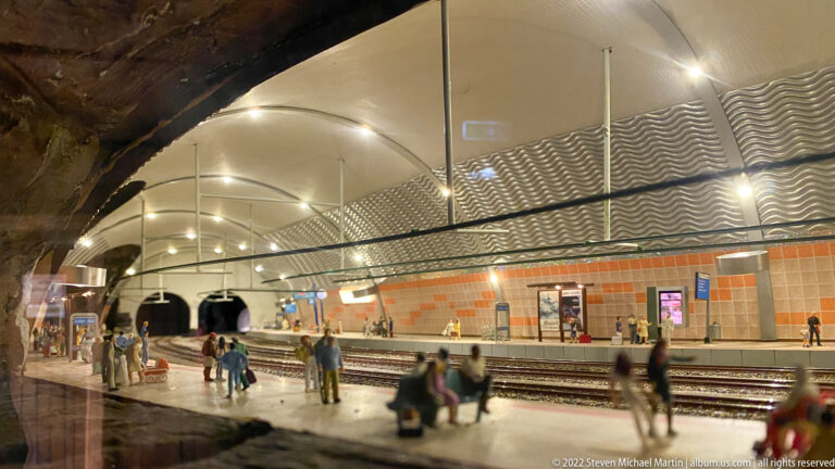 Miniature underground railway platform with passengers, curved vaulted ceiling, orange wall panels, and tracks.