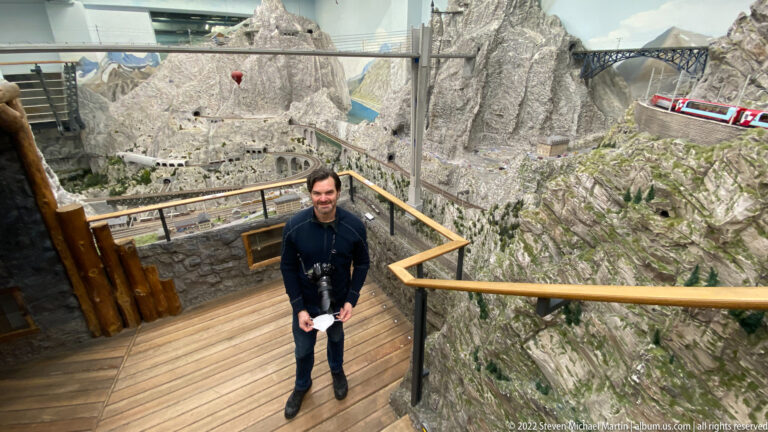 Visitor stands on a wooden platform overlooking miniature alpine mountains, railway bridges, tunnels, and trains.