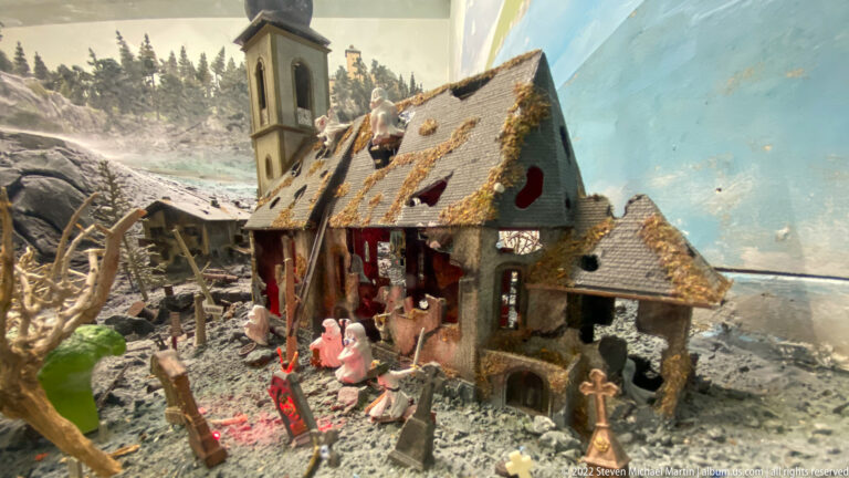 Weathered miniature alpine farmhouse with steep roof, rough stone walls, and tiny figures gathered around the entrance.