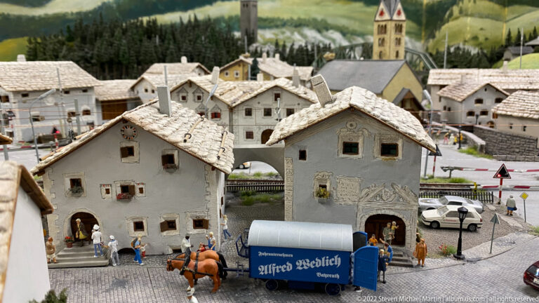 Blue horse-drawn Alfred Wedlich wagon passes through miniature alpine village with stucco houses and church tower.