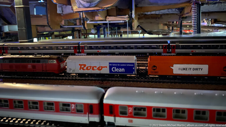 Model freight trains with “Roar Clean” and “I LIKE IT DIRTY” advertising pass behind red-and-white passenger coaches.
