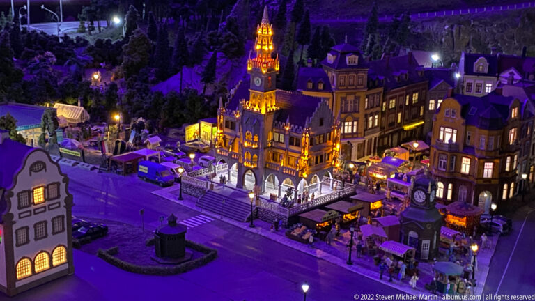 Illuminated miniature historic town square with ornate town hall, gabled buildings, cafés, pedestrians, and vehicles at night.