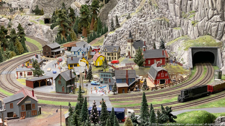 Miniature alpine village with church, colorful houses, looping railway tracks, station buildings, and mountain tunnel.
