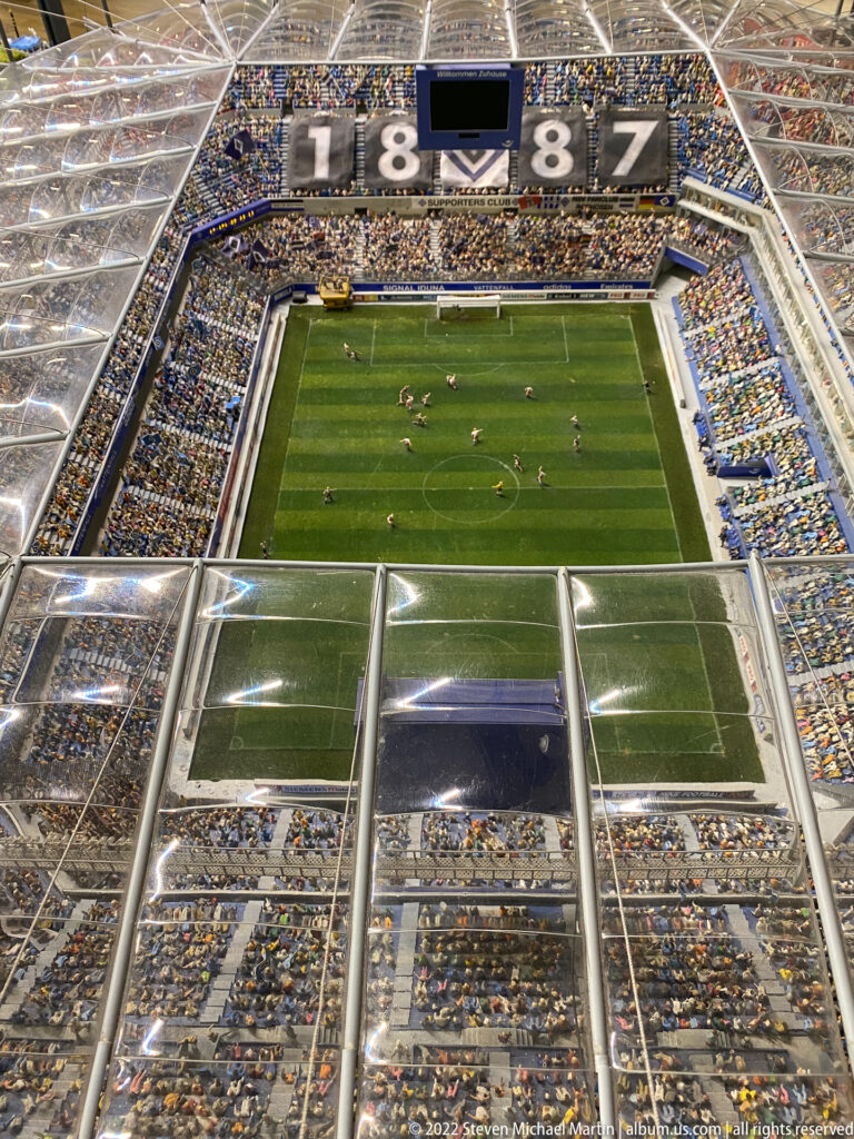 Miniature football match inside a large packed stadium with striped green pitch, players, roof structure, and scoreboard.
