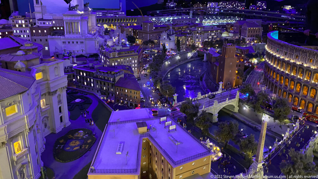 Wide miniature city-center panorama at night with illuminated civic buildings, roads, traffic, plazas, and an oval monument.