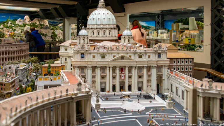 Miniature St. Peter’s Basilica facing a colonnaded square with central obelisk and tiny figures.