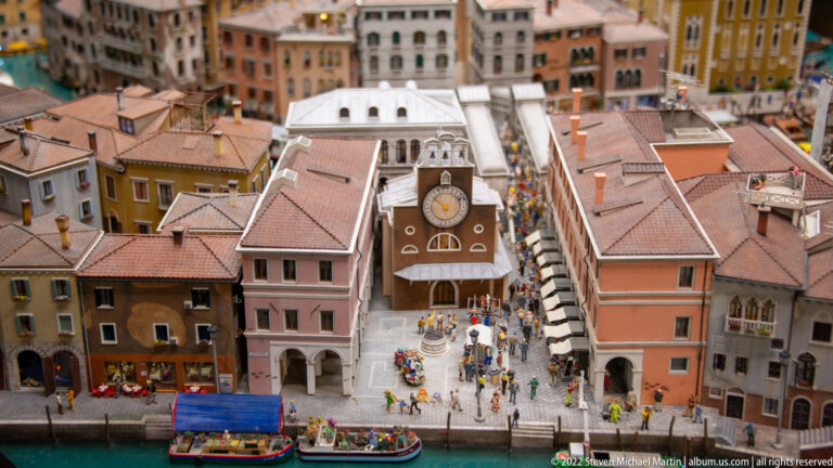 Miniature Venetian waterfront square with clock façade, crowded quay, colorful buildings, and passenger boats.