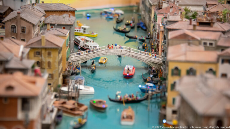 White arched pedestrian bridge crowded with figures above miniature gondolas and boats on a Venetian canal.