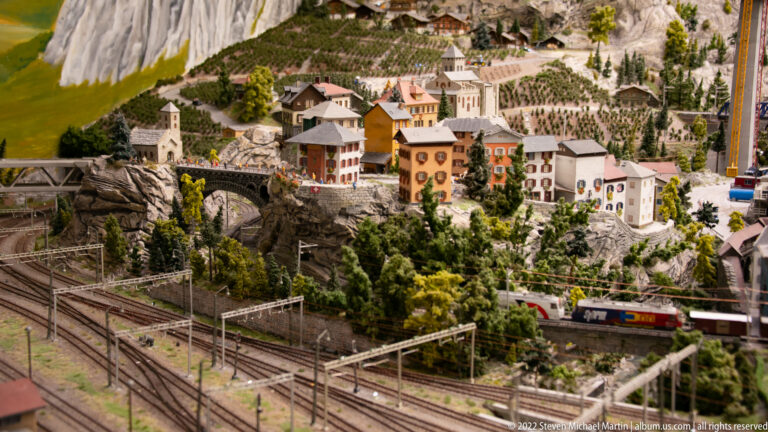 Miniature Alpine village above multiple electrified railway tracks, tunnels, bridges, and a passing train.