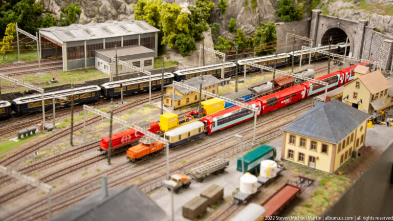Miniature rail yard with red passenger trains, dark coaches, freight equipment, service vehicles, and overhead wires.