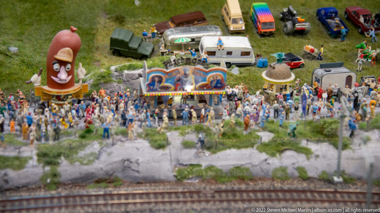 Crowded miniature festival field with caravans, parked cars, novelty figures, colorful stalls, and railway tracks in front.