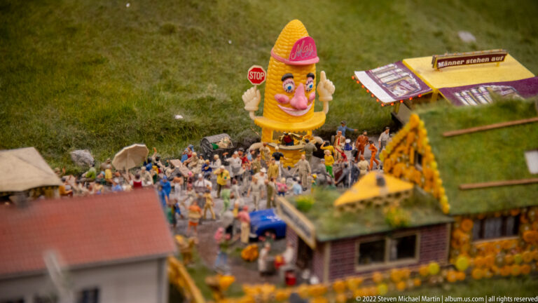 Miniature crowd gathered around a giant smiling corn figure beside tents, signs, and small roadside buildings.
