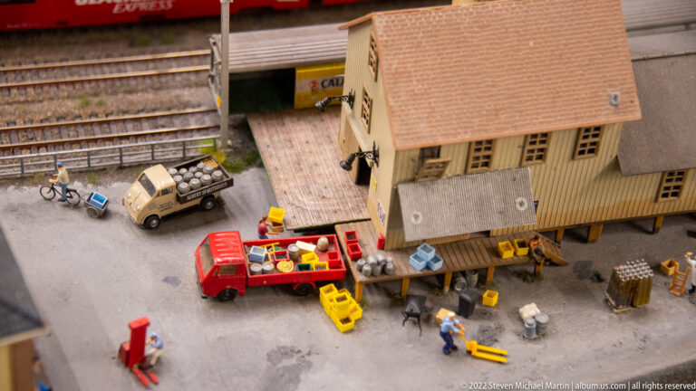 Miniature warehouse yard with red delivery truck, utility vehicle, workers, crates, carts, and railway tracks behind.