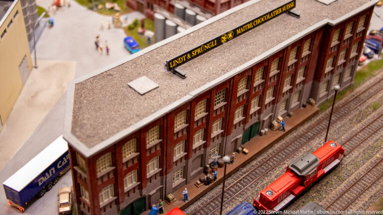 Miniature Lindt & Sprüngli brick factory beside railway tracks with a red train passing.
