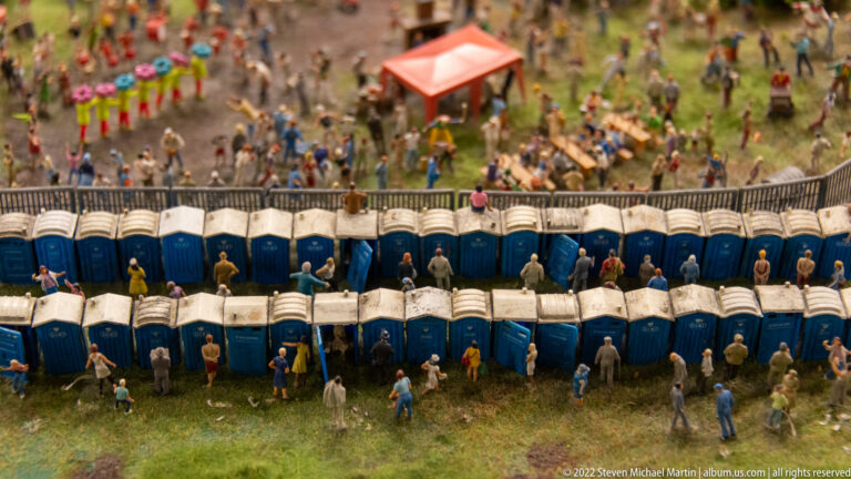 Rows of blue miniature portable toilets with many tiny figures at a crowded outdoor event.